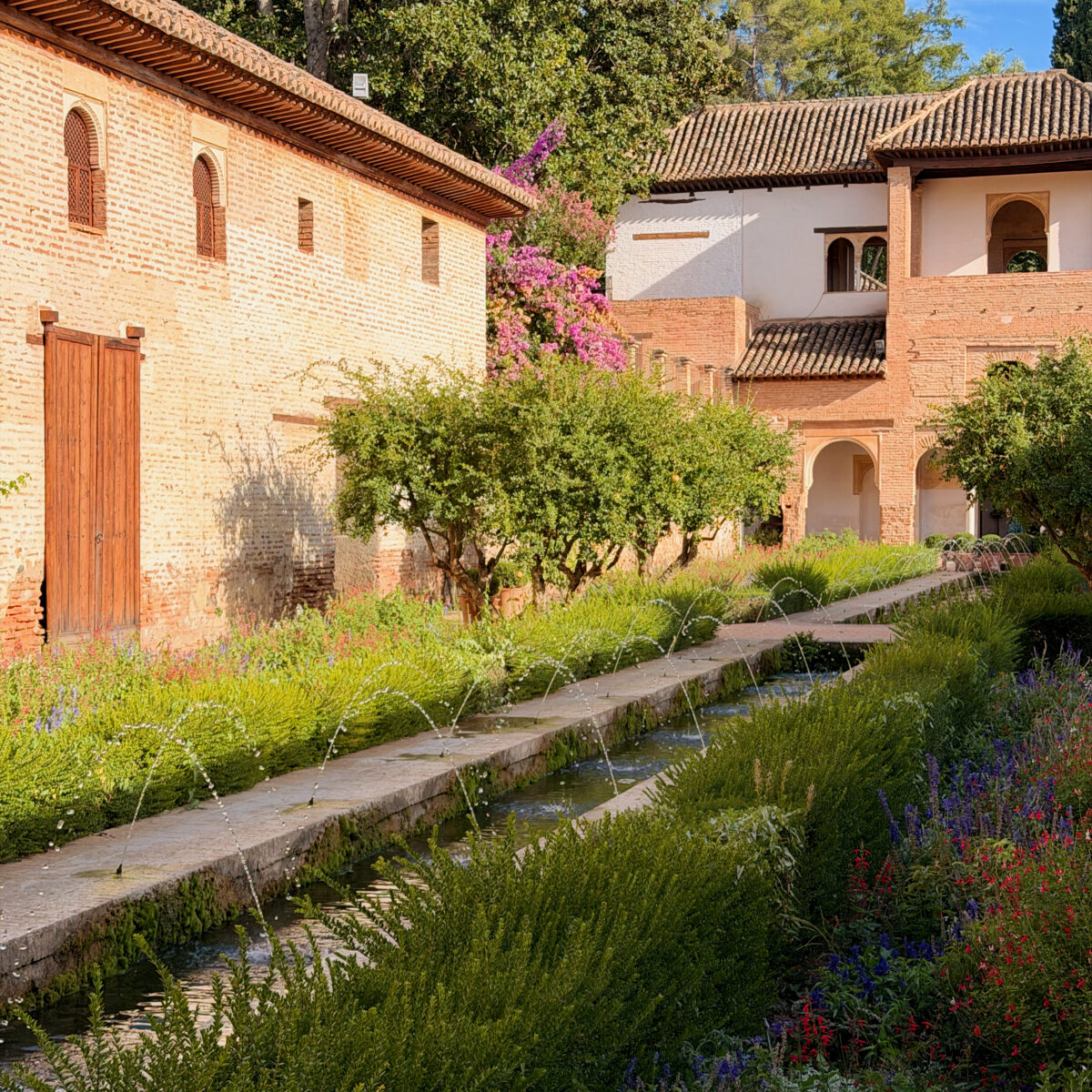 Brick buildings with tiled roofs line a narrow canal bordered by lush greenery and colorful flowering plants under a clear blue sky.