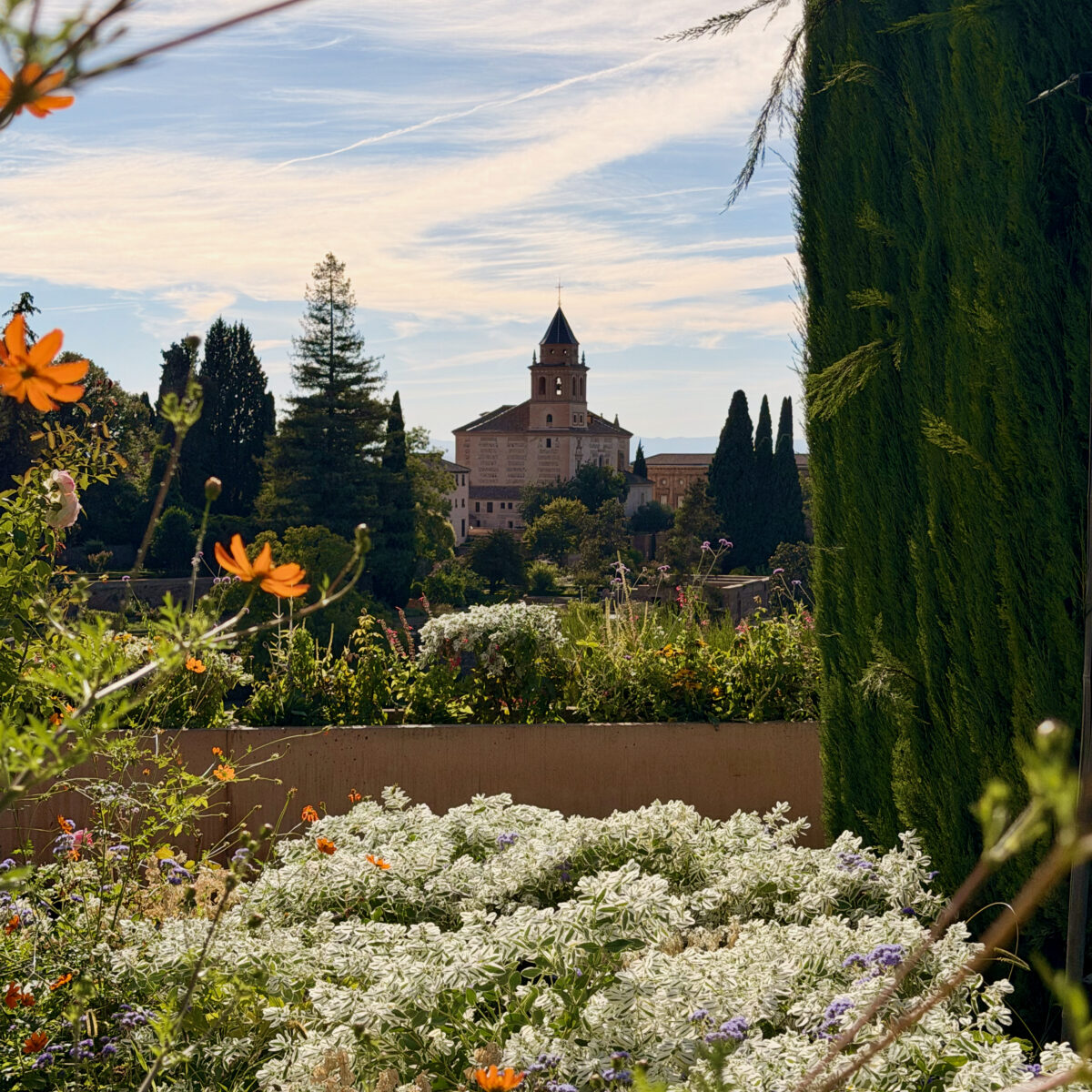 Colorful garden in the foreground with orange flowers and silvery leafy plants, a brick church and tall trees visible beyond under blue sky.
