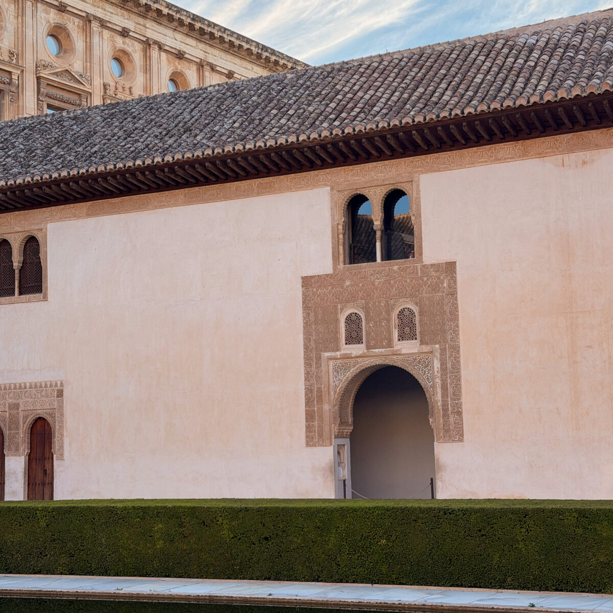 Clerestory windows and ornate archway carved into a beige wall, with a tiled roof and manicured hedges in the foreground.