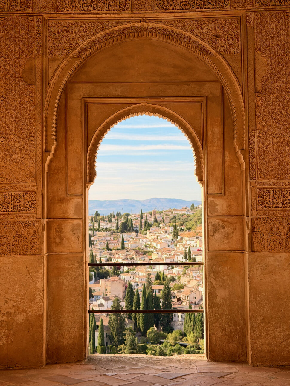 Intricately carved archway with ornate patterns frames a view of a sunlit hillside town dotted with trees and terracotta rooftops.