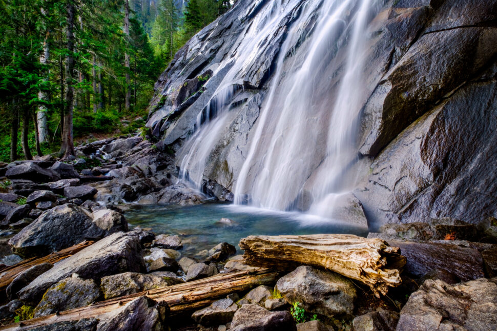 Waterfall cascading down a rocky cliff into a clear pool, surrounded by green trees and scattered boulders and logs.