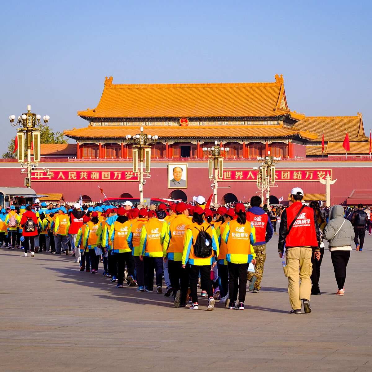 Long line of people wearing blue and yellow vests walking toward the Forbidden City’s Tiananmen Gate with red banners and flags in the background.