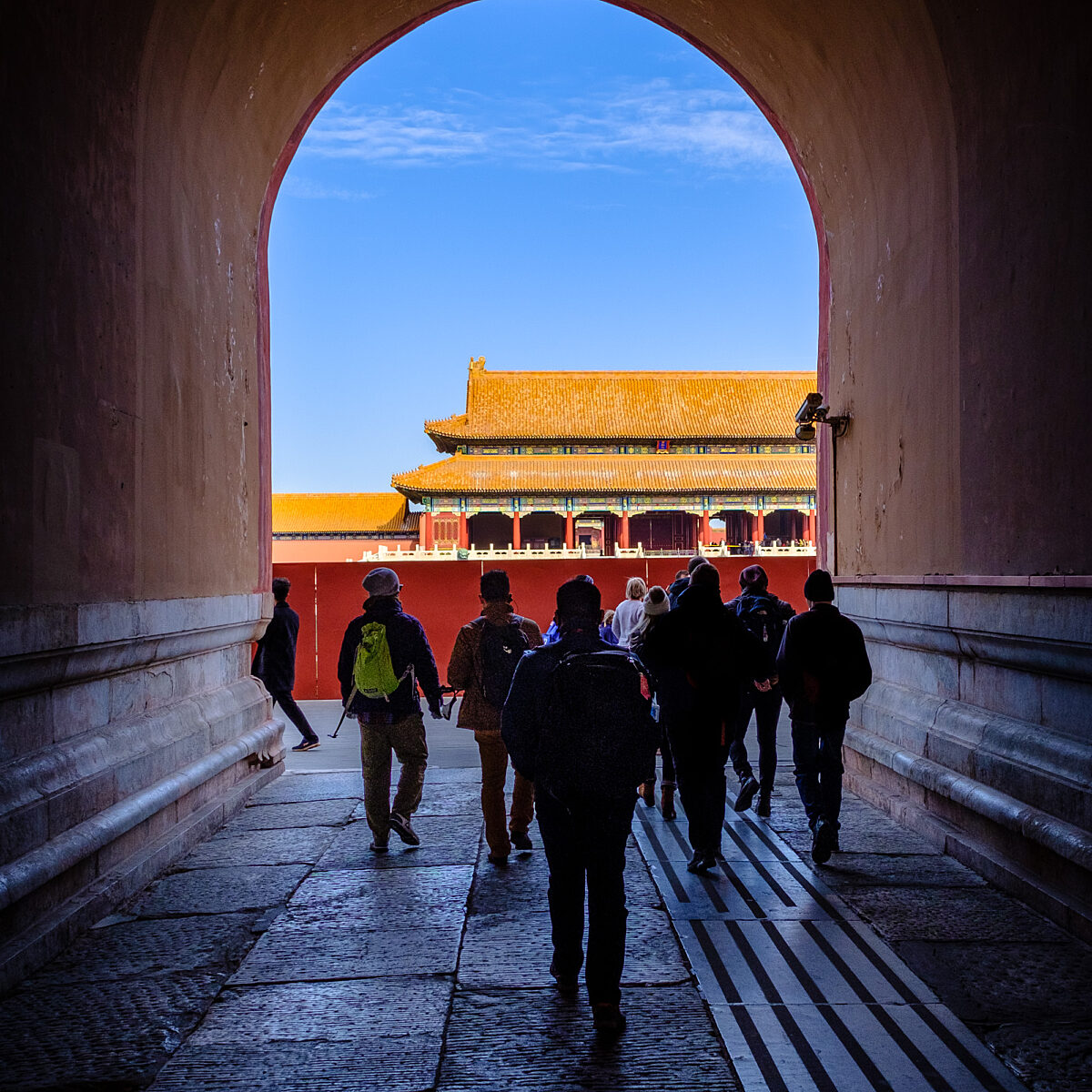 People walk through a dark arched tunnel toward a sunlit traditional Asian building with orange-tiled roof and red walls.