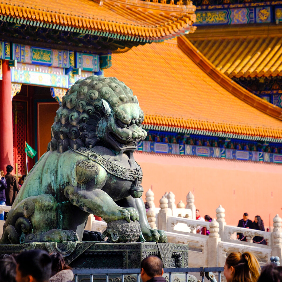 Bronze-green Chinese guardian lion statue with curled mane sits on a stone pedestal in a traditional temple courtyard, ornate orange-tiled roof and colorful decorative eaves in the background.