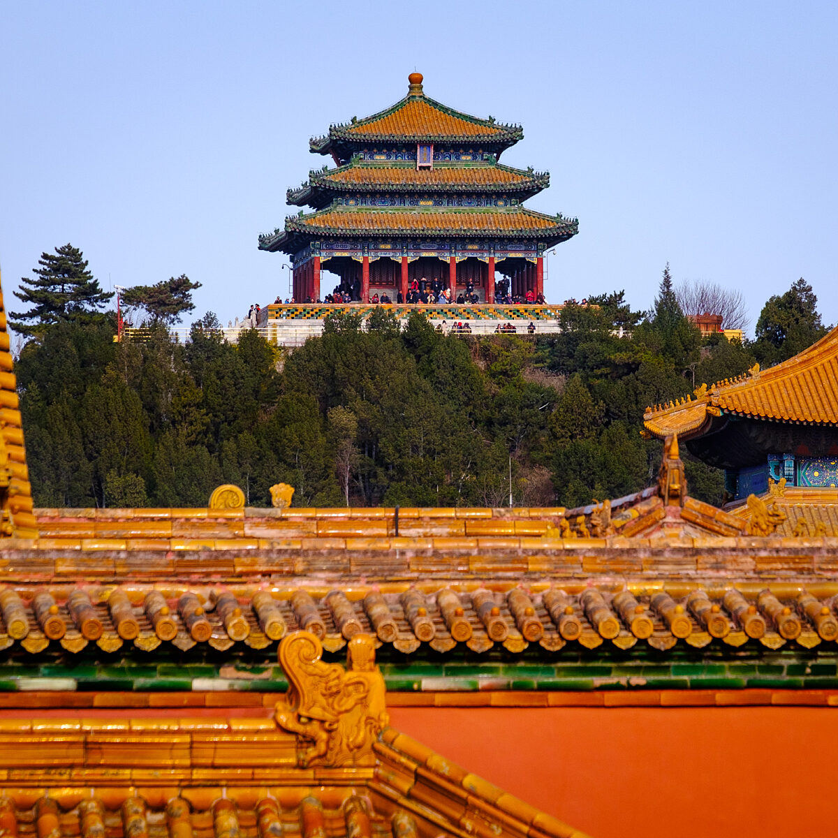 Golden-tiled rooftops with ornate dragon motifs frame a multi-tiered green-and-gold pagoda atop a hill, surrounded by trees against a clear blue sky.