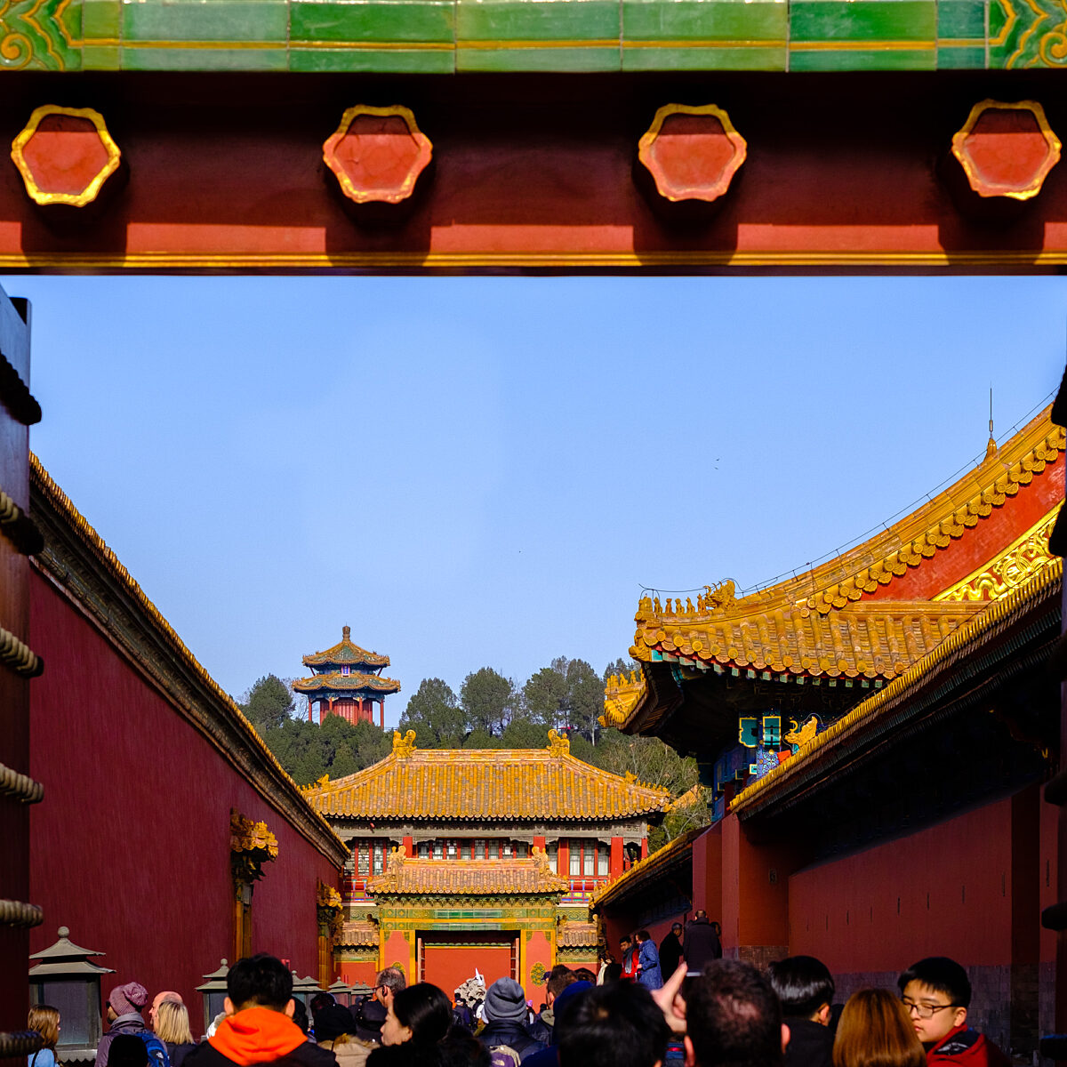 Vivid Chinese temple complex framed by a colorful doorway, with ornate yellow-tiled roofs and a blue sky visible above crowds of visitors.