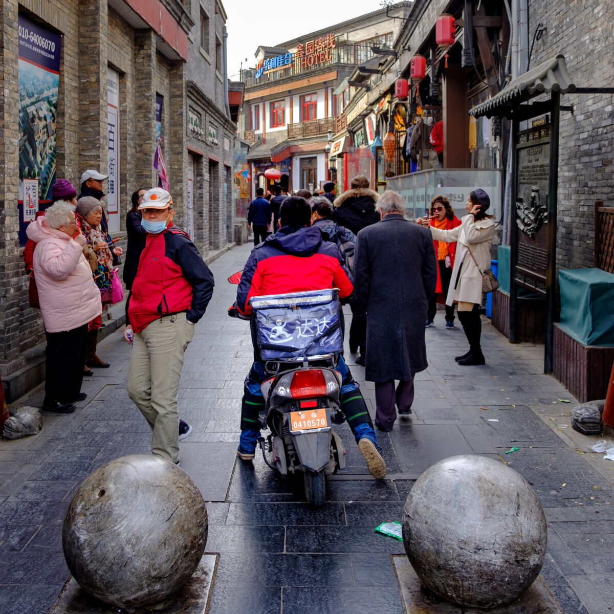 Bustling narrow street lined with shops and pedestrians; a person on a scooter pushes through while others chat and stroll along slick stone pavement.