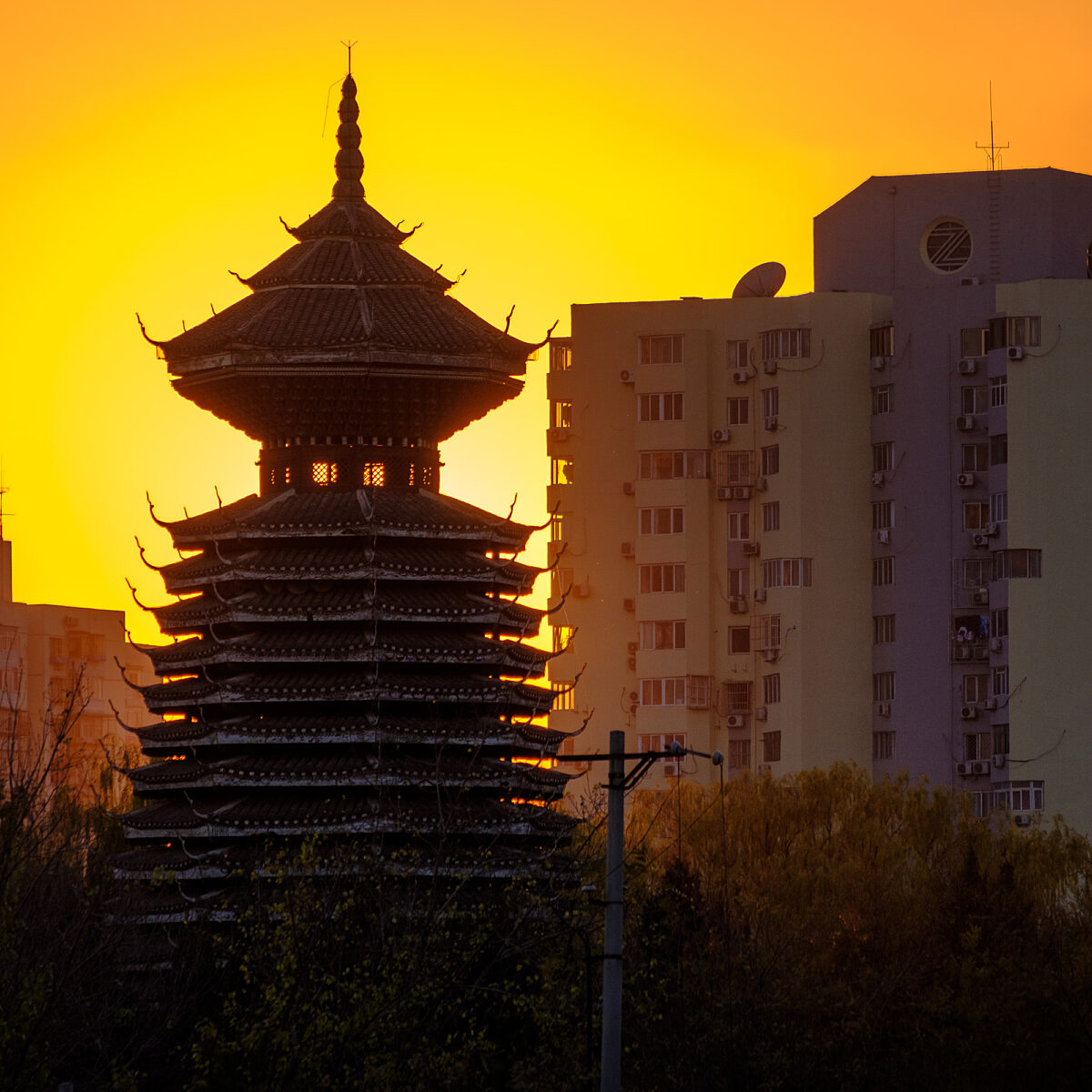 A multi-tiered, ornate pagoda stands among modern high-rise buildings against a bright orange sunset.