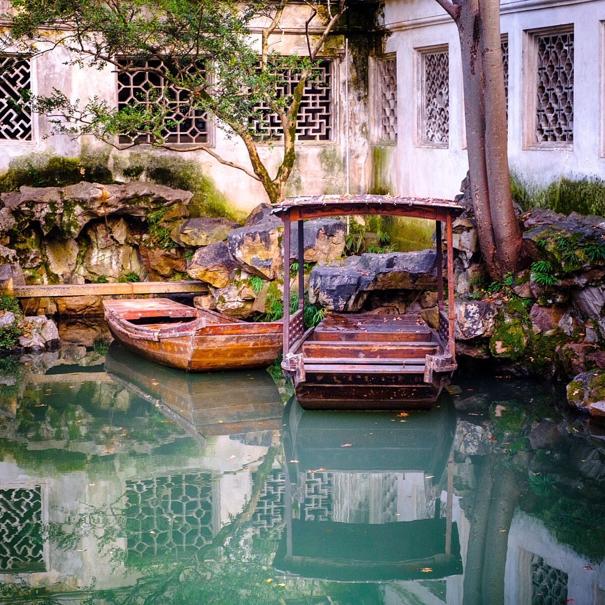 Two wooden boats float on calm green water beside a mossy stone wall and a white-walled building with lattice windows.