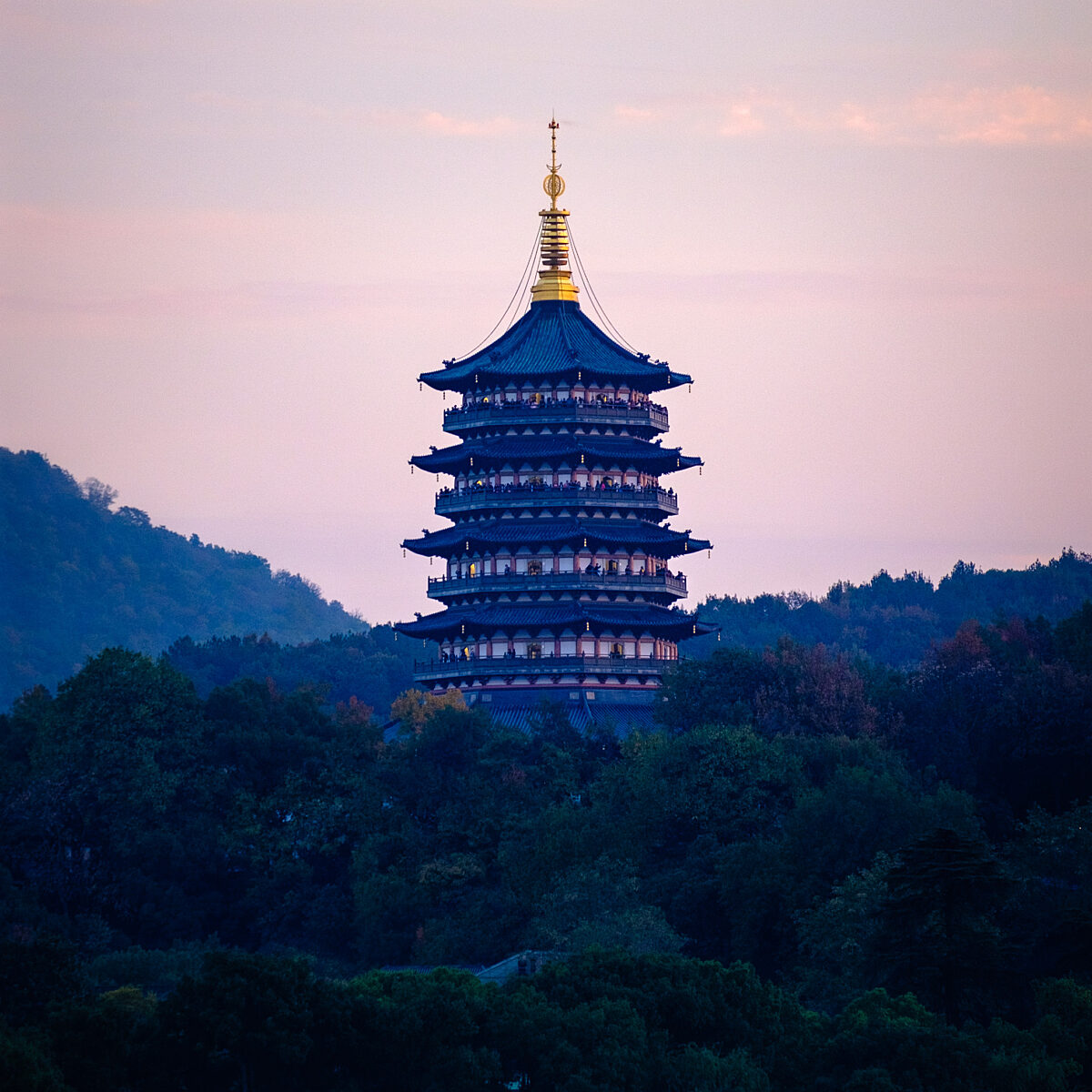 Nine-tier pagoda with blue roof rises among trees near a water body at dusk, gold spire and pink sky in background.