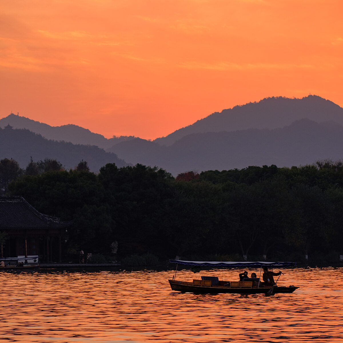 Sunset over a lake with layered silhouettes of mountains and a small boat carrying people on calm, orange-tinted waters.