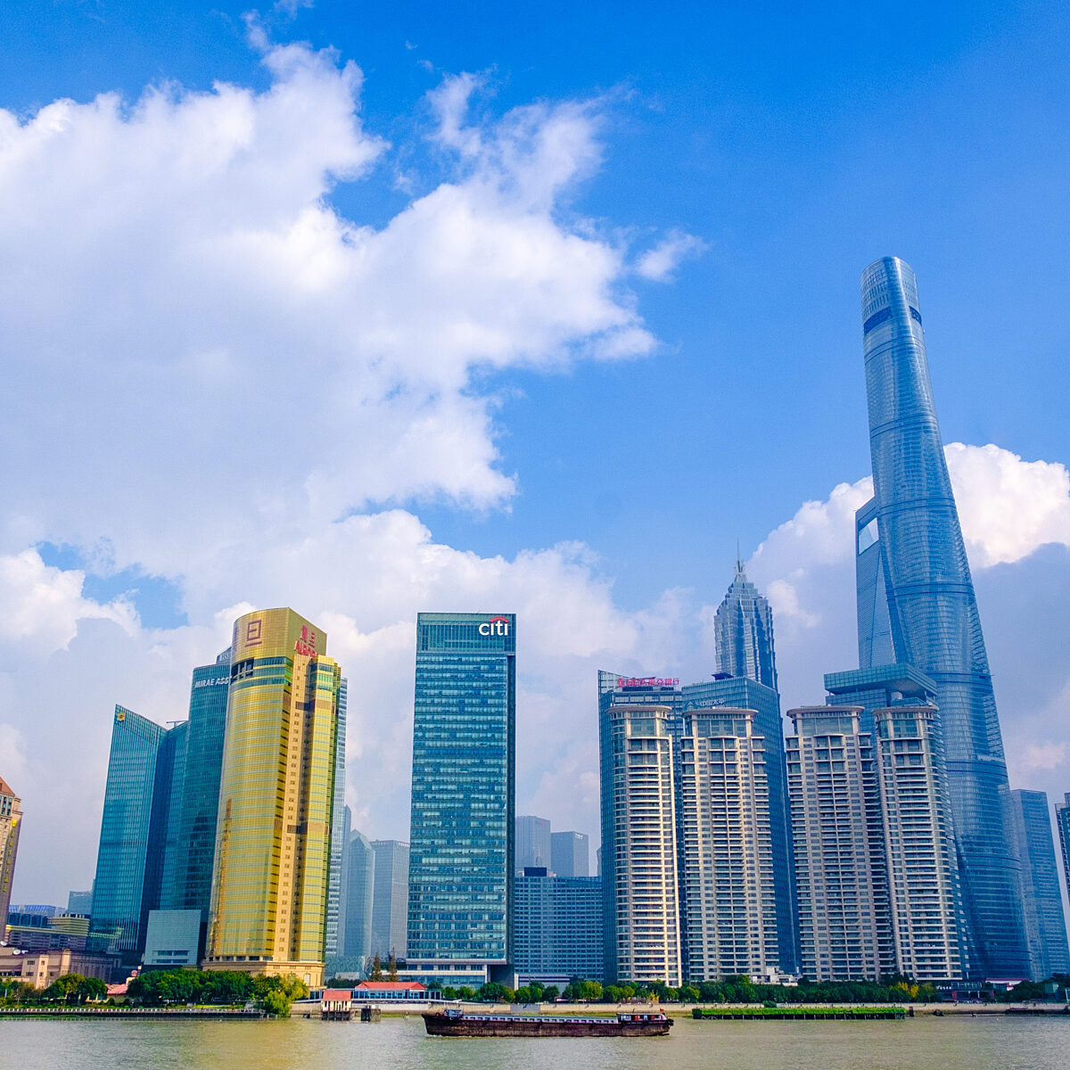 Skyscrapers line a riverfront under a bright blue sky with scattered white clouds, including a tall, twisting tower on the left and a slender, modern high-rise on the right.