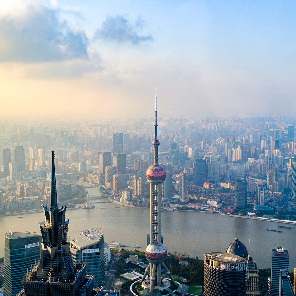 Aerial view of Shanghai with the Oriental Pearl Tower, tall skyscrapers, and the Huangpu River winding through a dense cityscape under a hazy, sunlit sky.