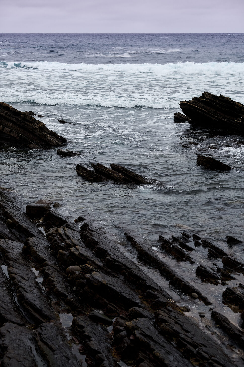 Layers of dark, jagged rocks jut into a choppy sea with white-capped waves under a cloudy sky.