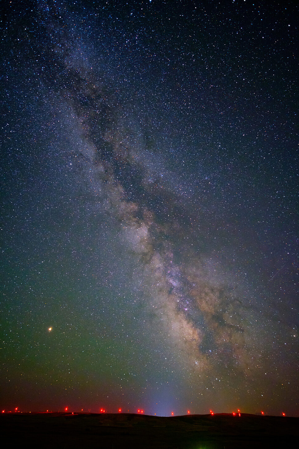 Milky Way stretches diagonally across a clear night sky filled with stars, with a faint greenish horizon and small red lights along the distant ground.