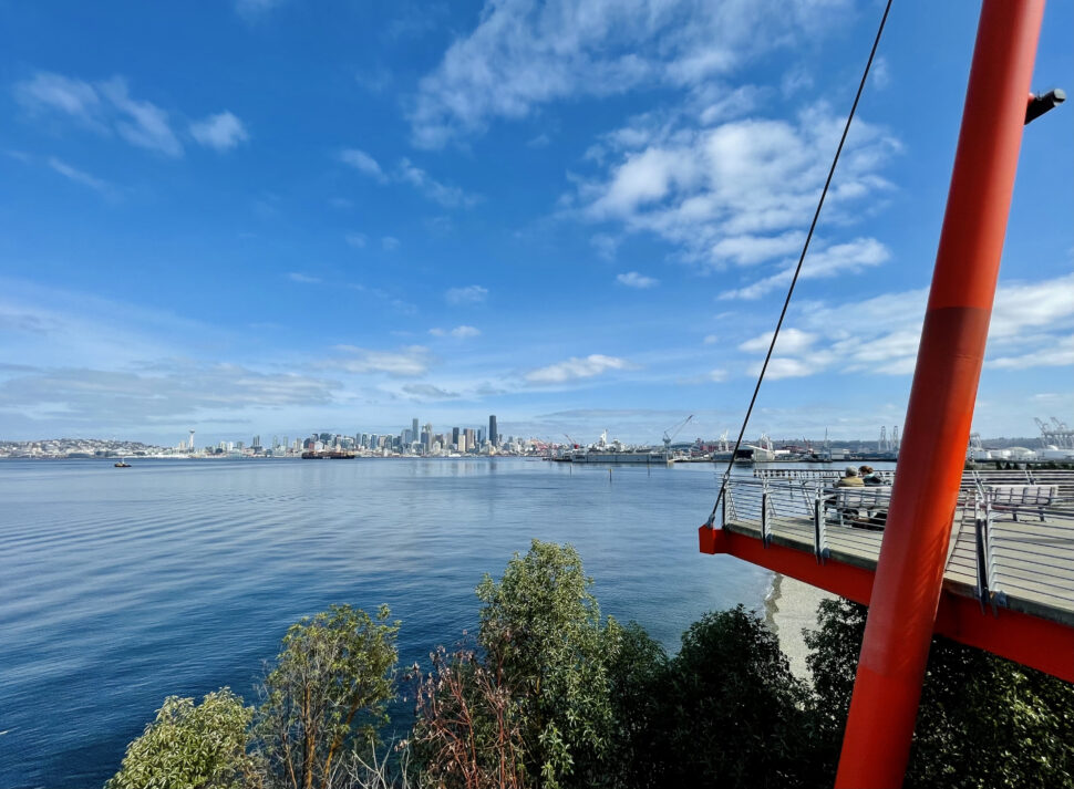 Sturdy red framework and railing extend over blue water, with a skyline of tall buildings across the harbor under a partly cloudy sky.
