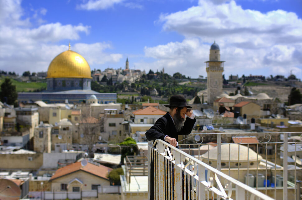 A bearded man in a black suit and hat stands on a balcony in front of a cityscape with the golden Dome of the Rock and a tall minaret in the background.