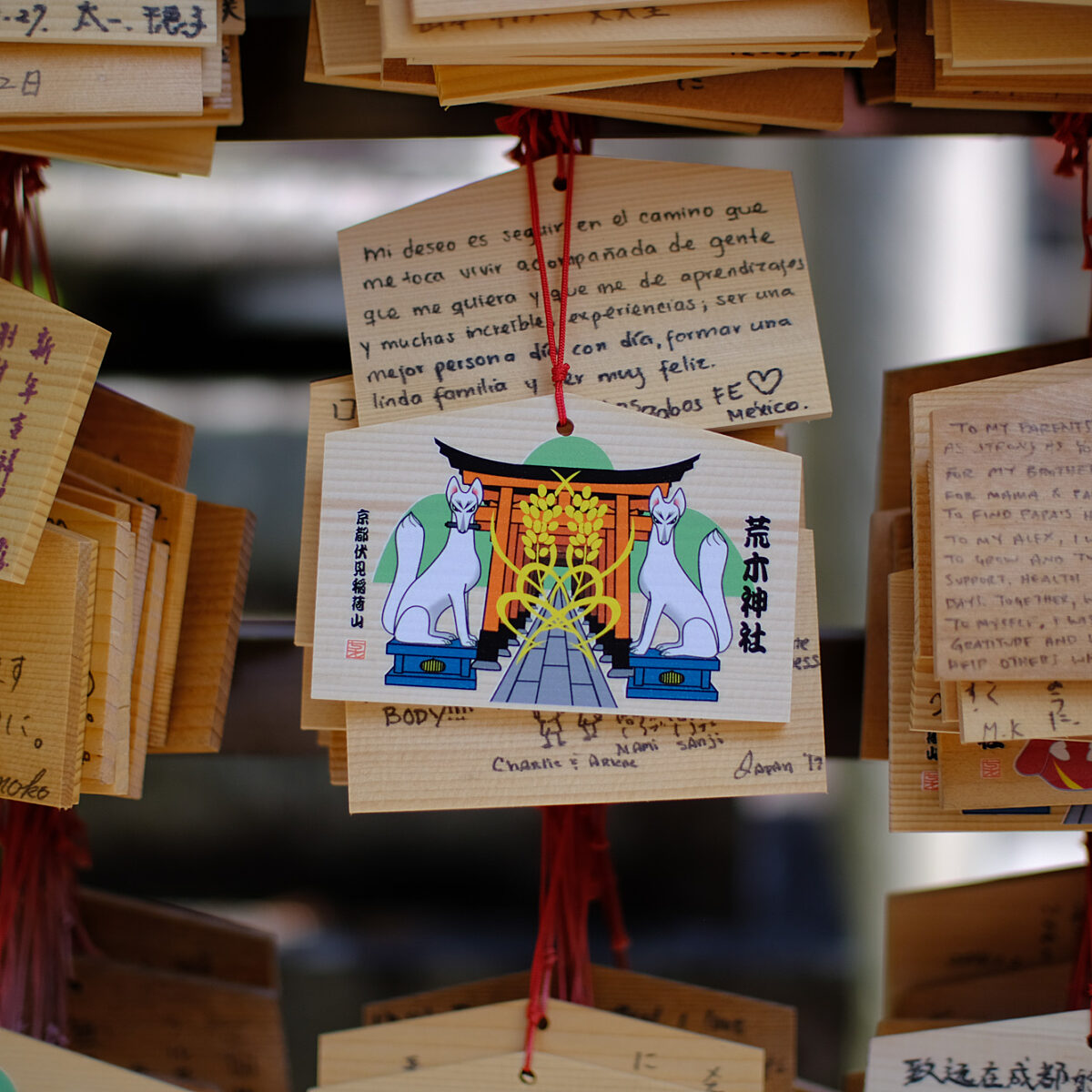 Wooden prayer plaques with handwritten messages in various languages hang from red cords, surrounding a central plaque featuring a shrine illustration with two fox statues.