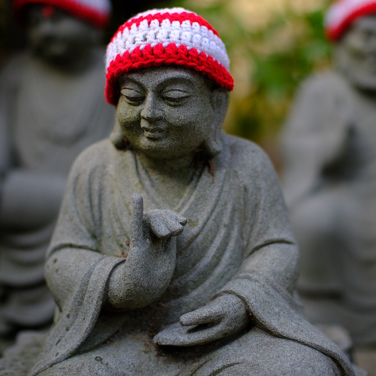 Stone statue of a meditating figure wearing a red-and-white knitted cap, with one hand raised and the other resting in lap.