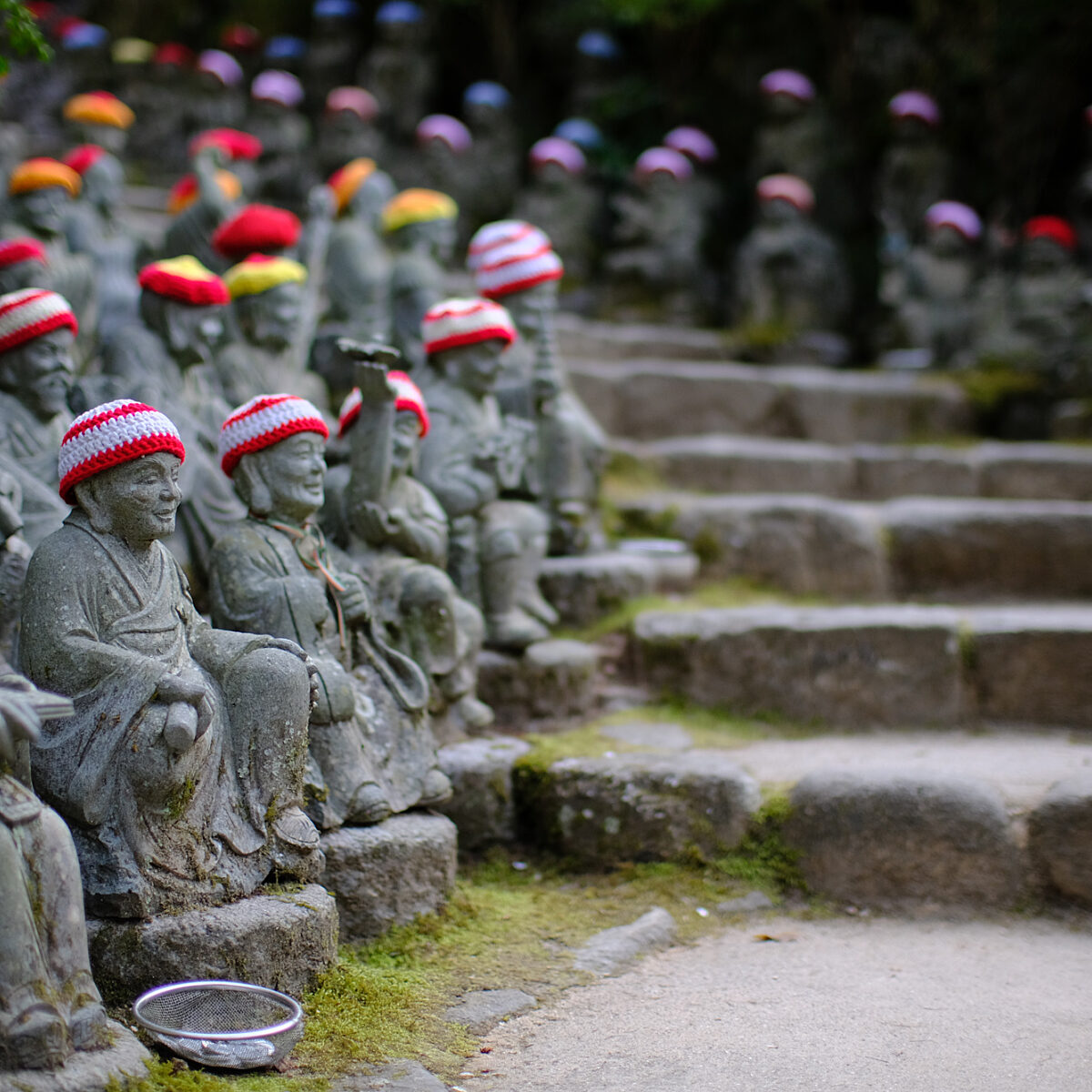 Row of stone statues wearing red and white knit hats along a mossy stone steps path in a garden.