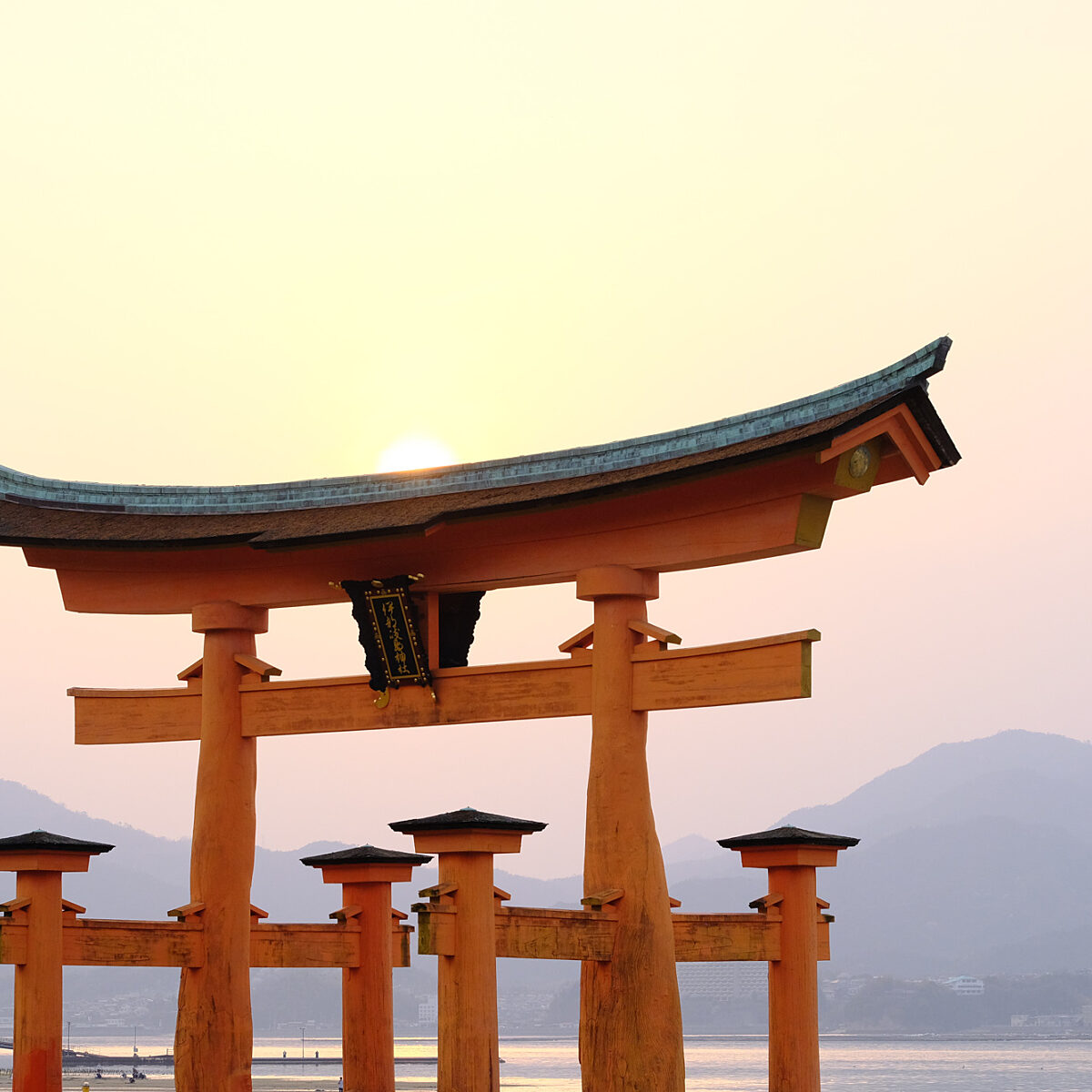 A bright orange torii gate rises over a calm body of water with distant mountains at sunset.
