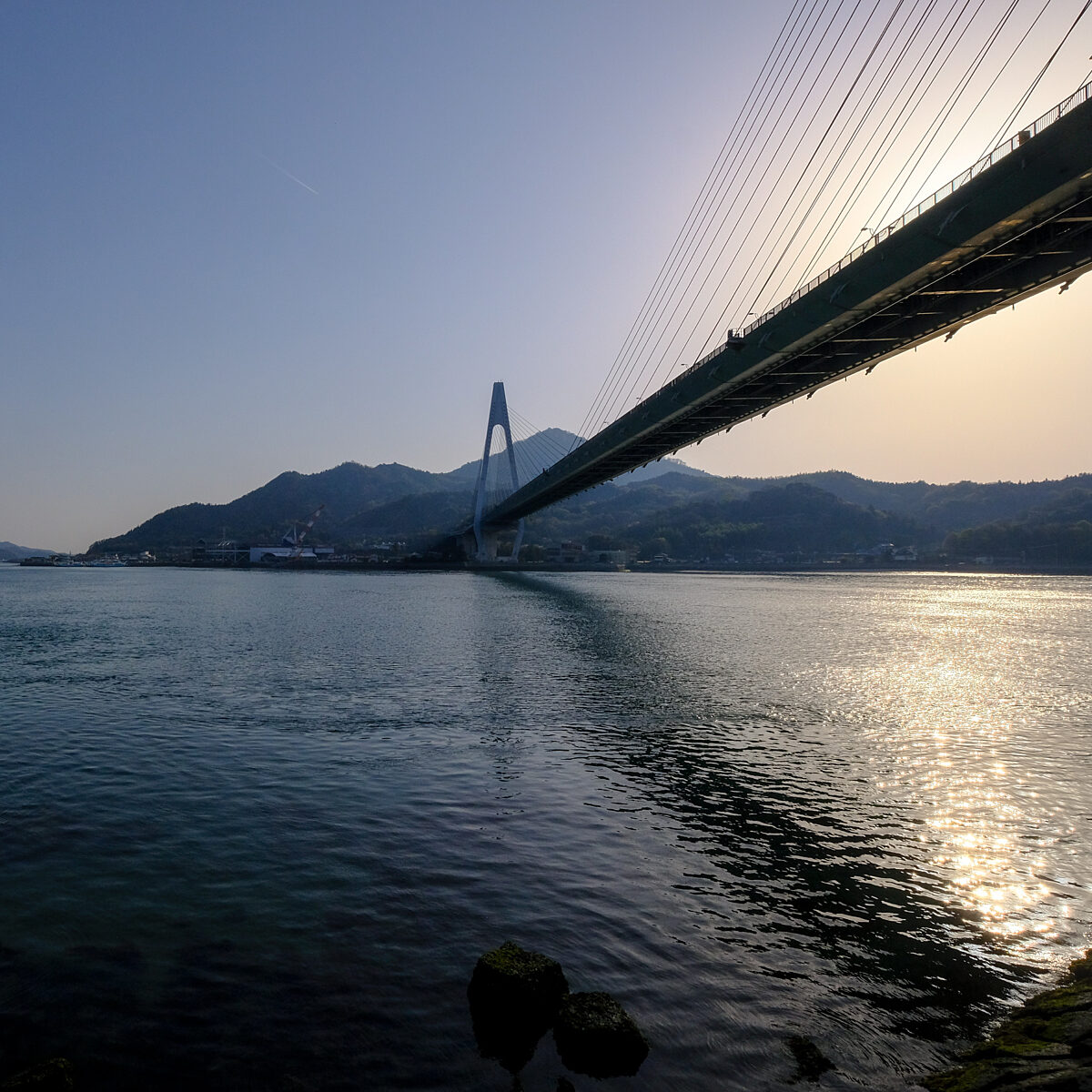 Cable-stayed bridge spans a calm body of water with hills in the background and sunlit reflections on the surface.