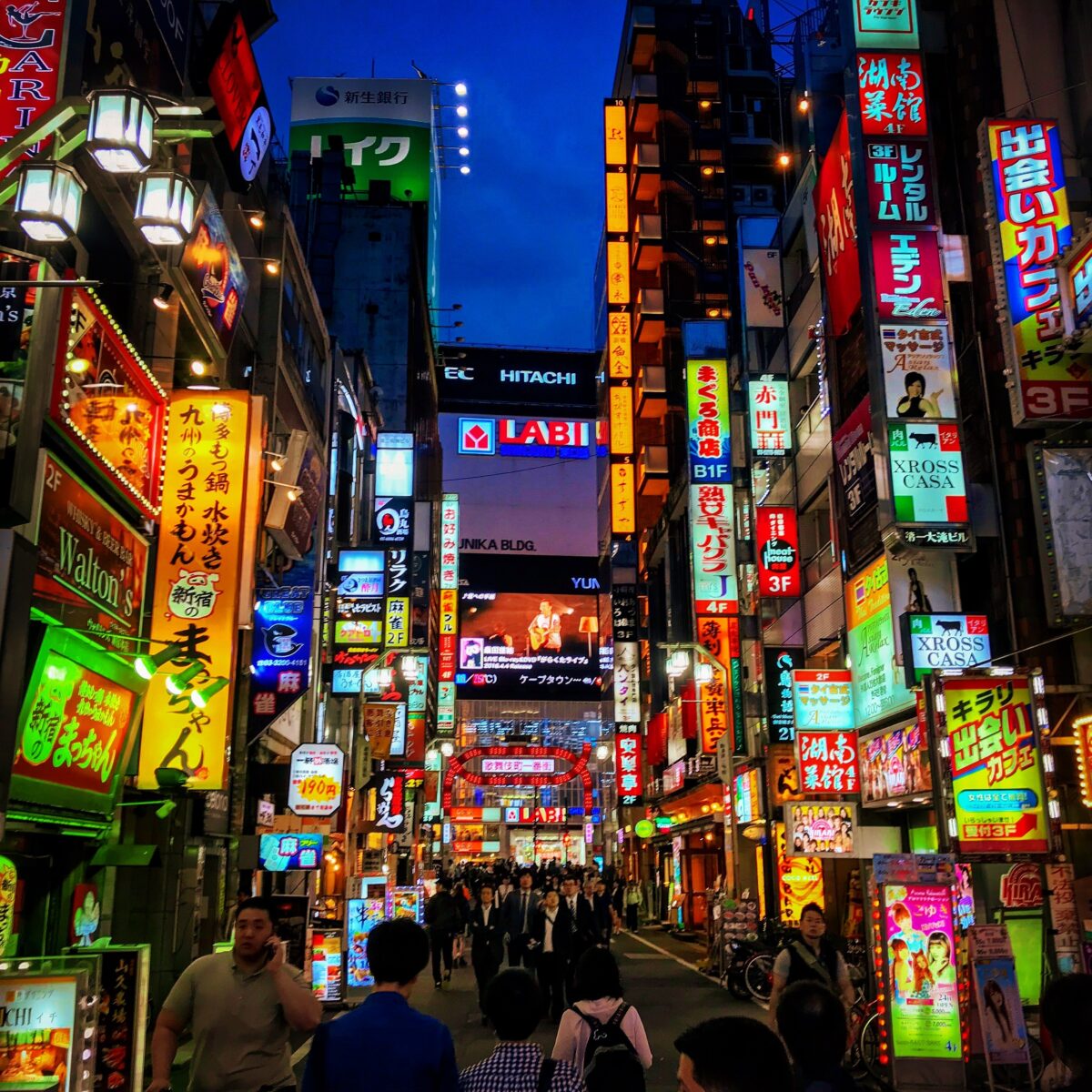 Bustling neon-lit street in a crowded city alley, with colorful signs in Japanese on tall buildings and pedestrians walking beneath the evening sky.