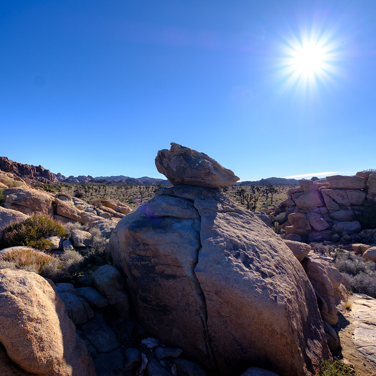 Desert rocky landscape under a bright sun, with large boulders in the foreground and sparse vegetation across a rugged terrain.