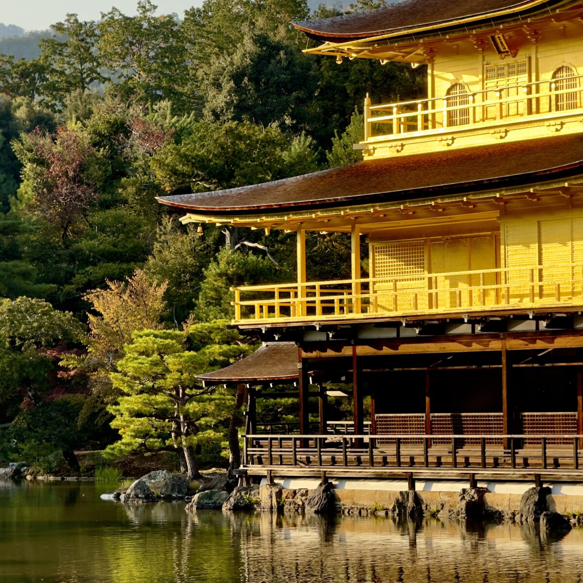 Golden, multi-tiered temple by a calm pond, with surrounding trees and rocky shoreline.