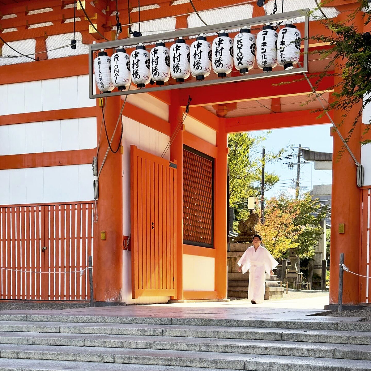 Stone steps lead to a bright orange temple gate with hanging white lanterns and a person in a white robe standing near the entrance.