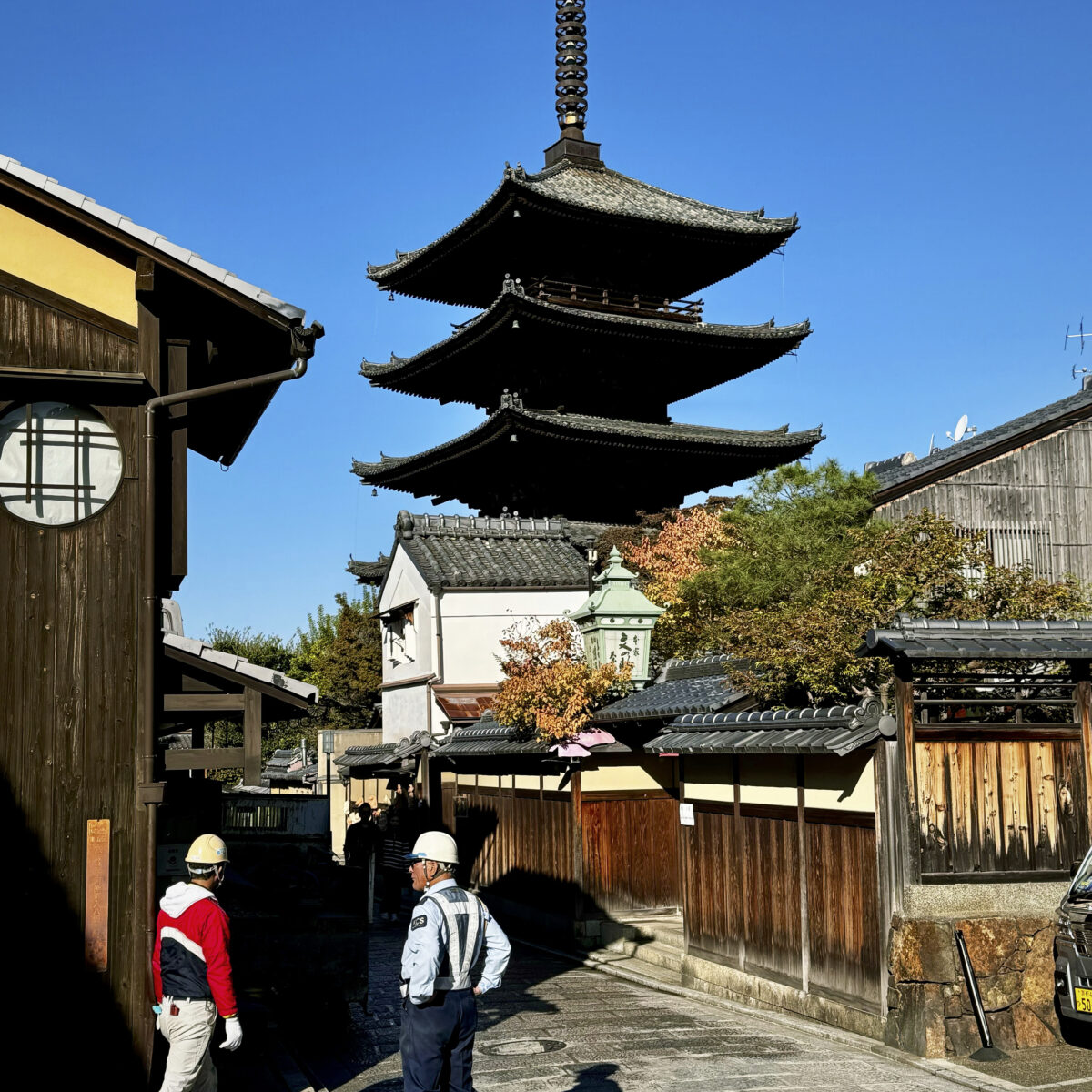 Five-story pagoda with a tall spire rises above a traditional Japanese street lined with wooden buildings and a stone-paved path.