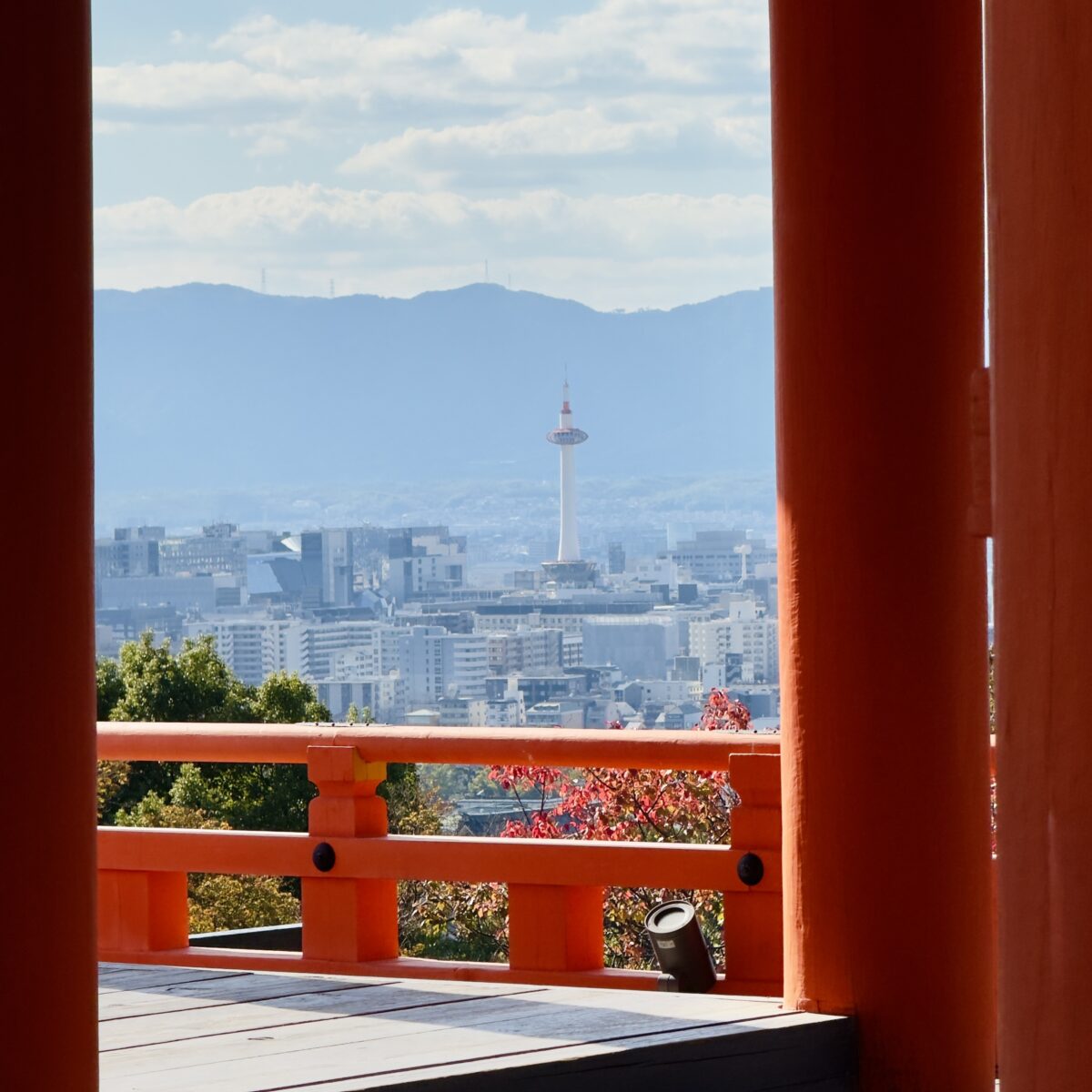 City skyline framed by orange temple railing and posts, with a tall tower and distant mountains under a blue sky.