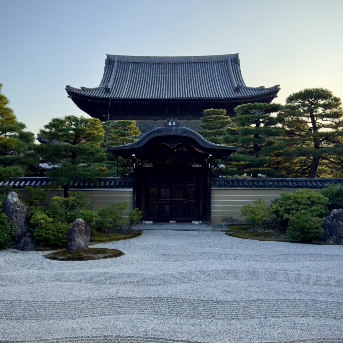 Stone gate and traditional Japanese building with curved roof, trimmed trees, and a gravel Zen garden in front.