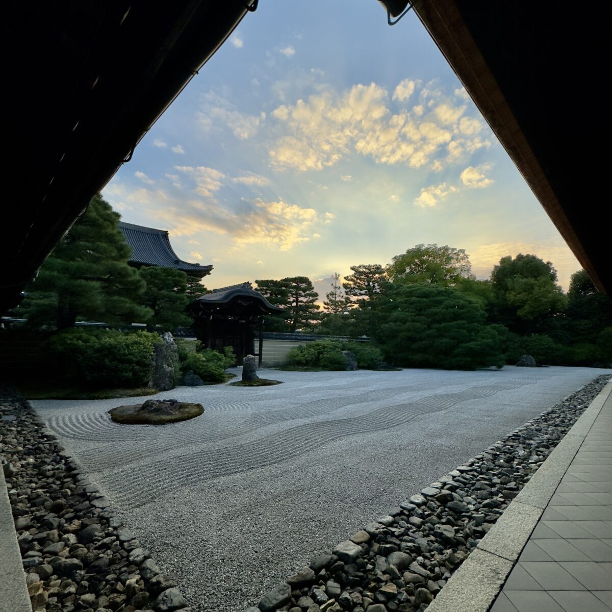 View from a framed courtyard to a Japanese rock garden, with raked gravel, stones, manicured trees, and a traditional roofed structure against a blue sky with clouds.
