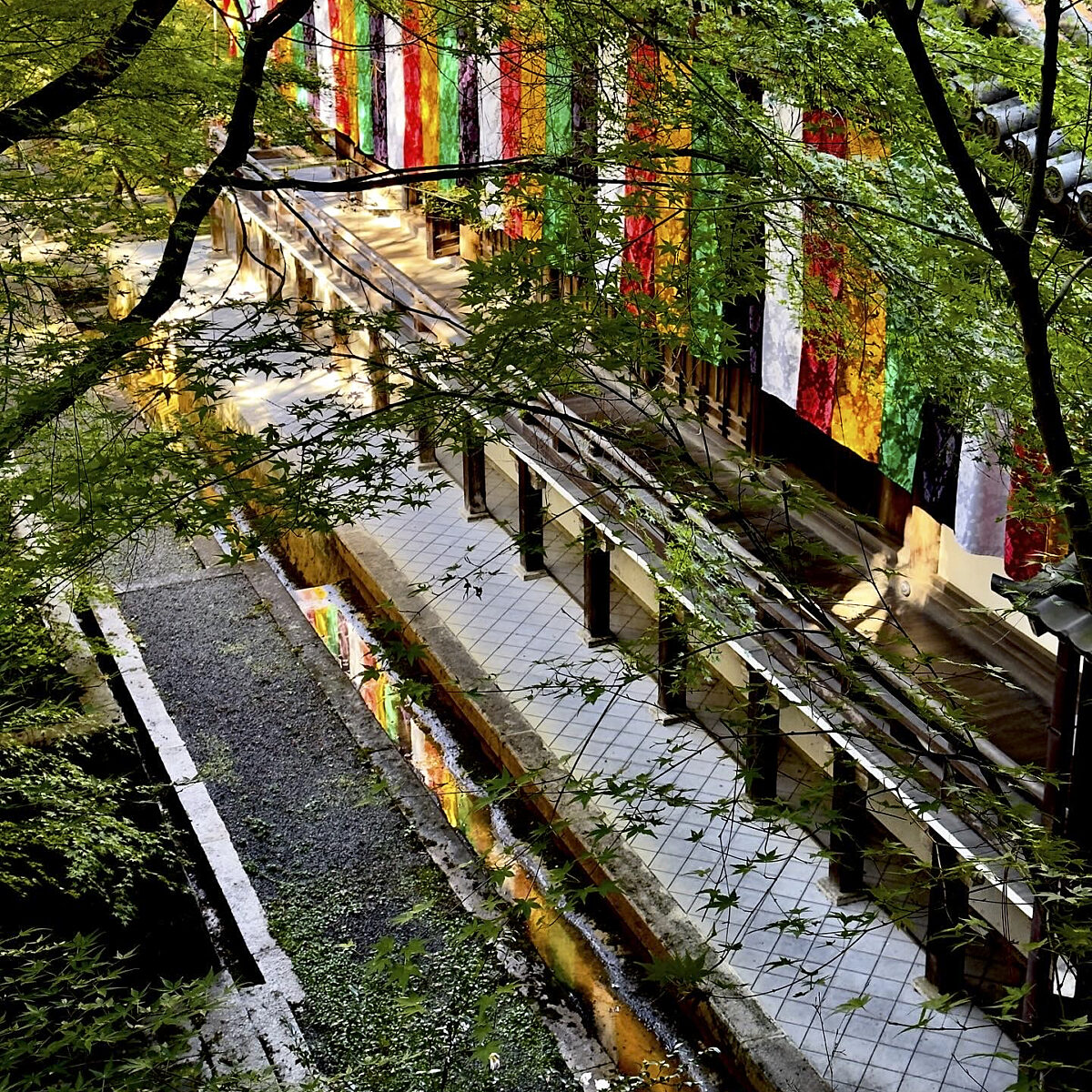 Colorful vertical banners hang along a walkway lined with green trees and railings, reflecting in a narrow water channel below.