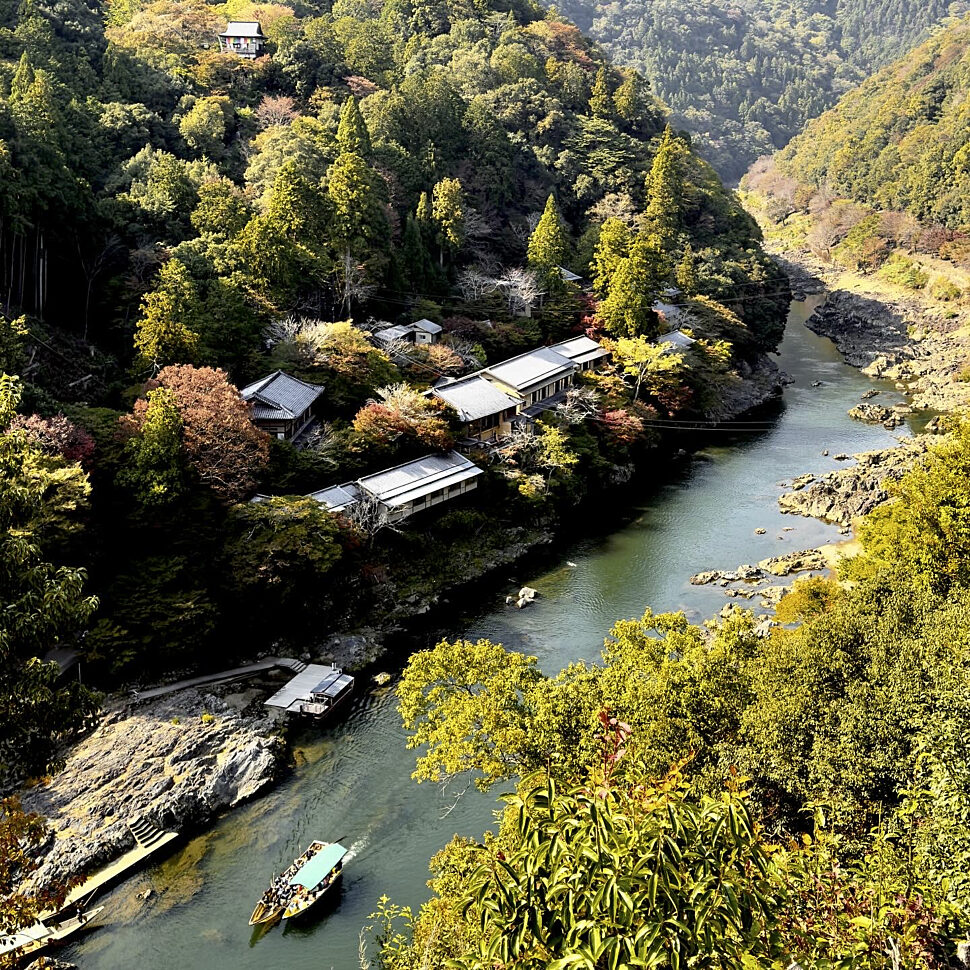 A riverside village with traditional buildings along a winding greenish river, surrounded by dense autumn trees on hillsides.