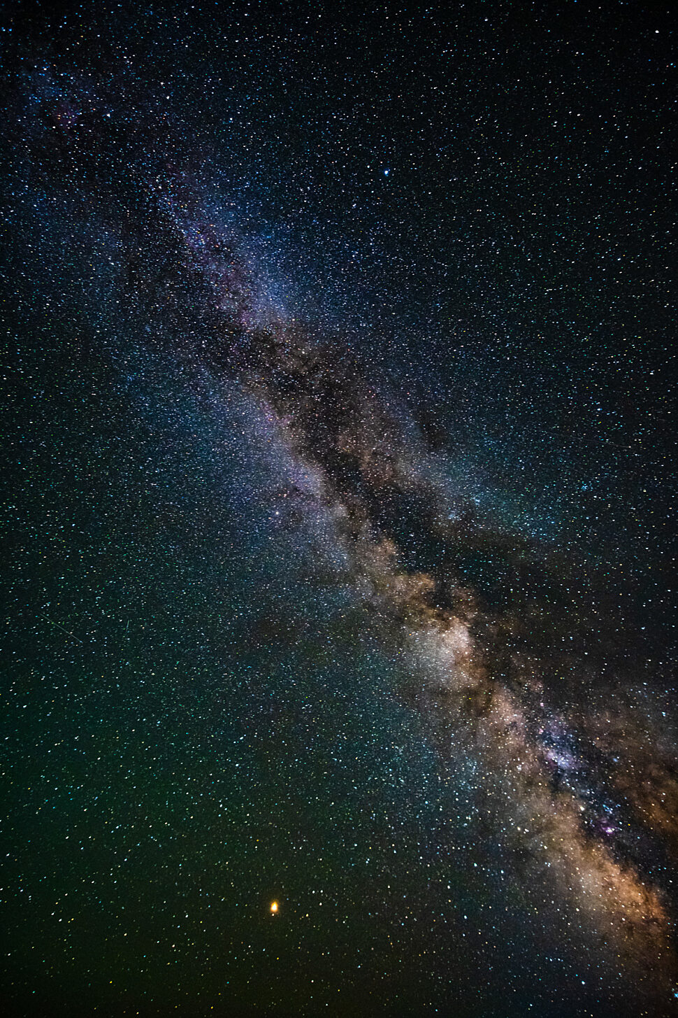 View of the Milky Way's bright central band across a dark, star-filled sky with a yellowish foreground object near bottom center.