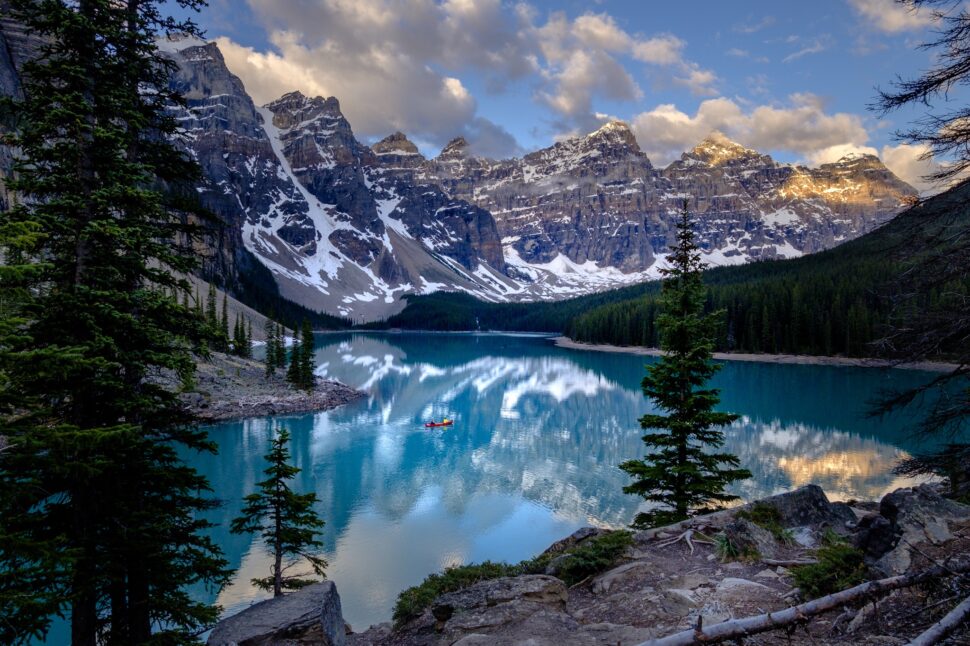 Turquoise lake reflects snow-dusted jagged mountains under a blue sky, with evergreen trees along rocky shore and a small red kayak on the water.