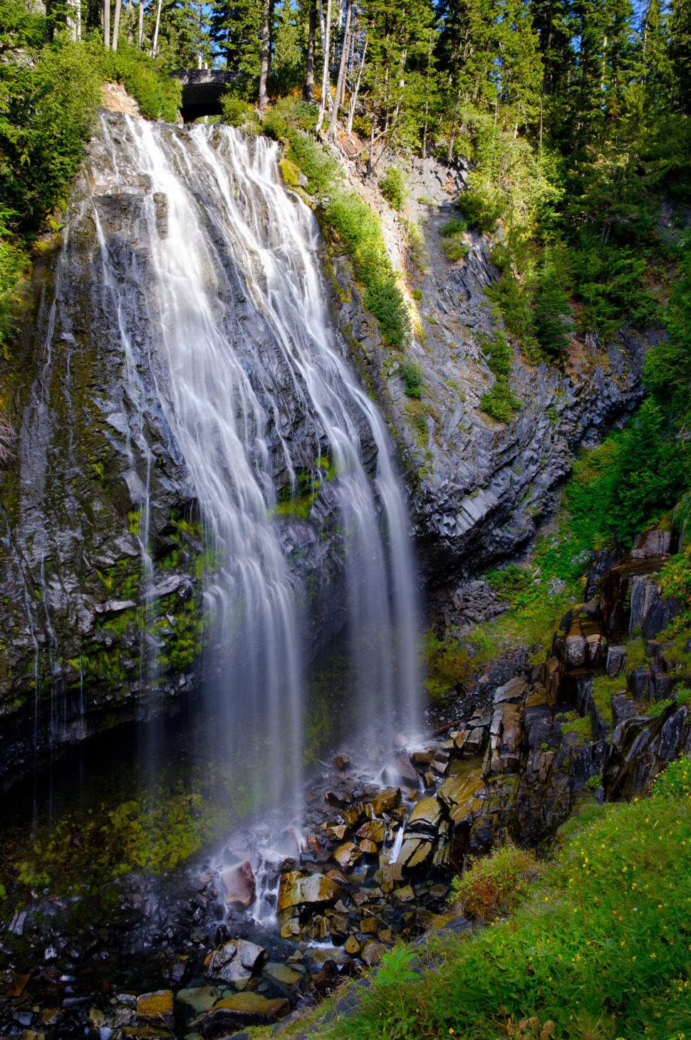 Tall multi-branch waterfall cascading down a rocky cliff surrounded by green trees and moss, with a rocky pool and scattered boulders at the base.