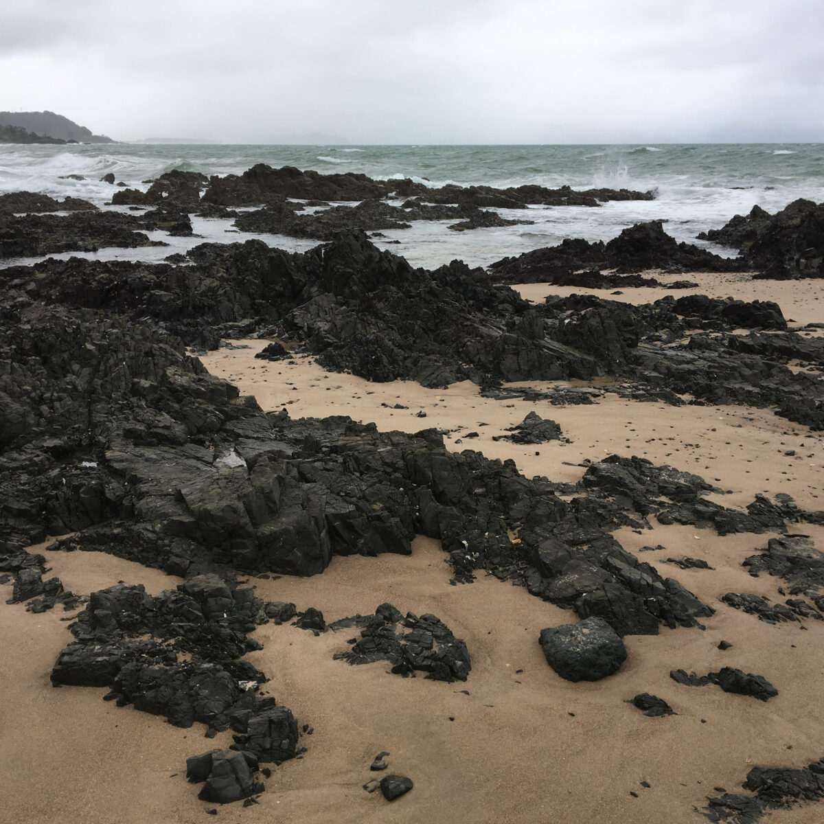Dark, jagged rocks scattered across a wet sandy beach with rough ocean waves and a gray, cloudy sky in the distance.