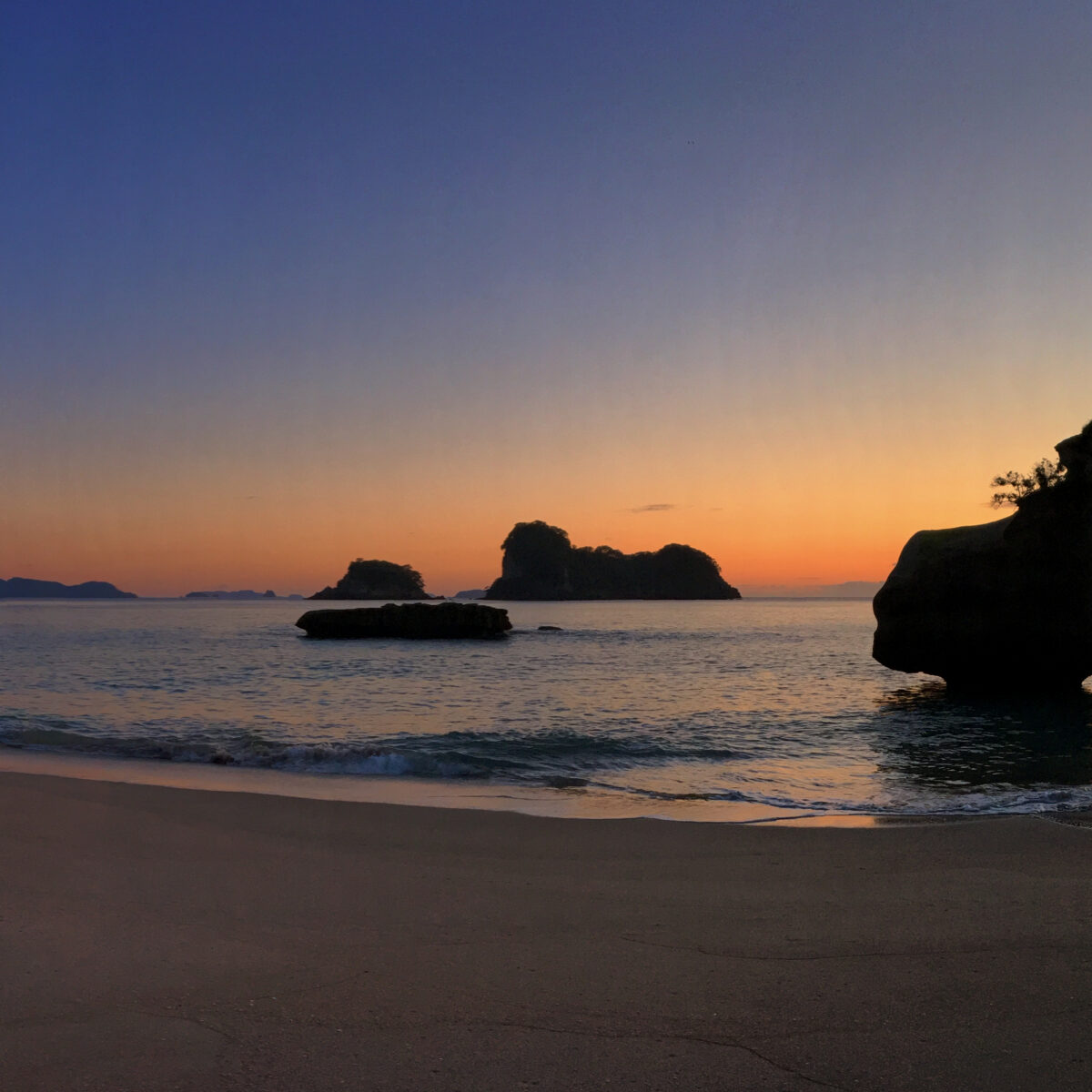 Sunset over a quiet bay with rocky formations, a sandy beach, and calm water; silhouettes of cliffs and trees frame the horizon as the sky fades from orange to deep blue.