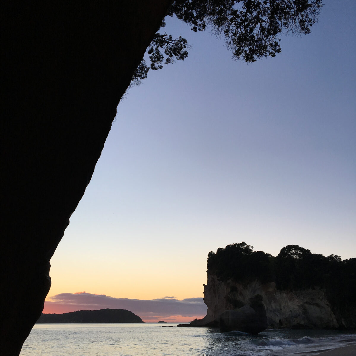 Sunset over calm sea framed by dark cliff faces and silhouettes of distant hills, with gentle waves lapping at a rocky shore.