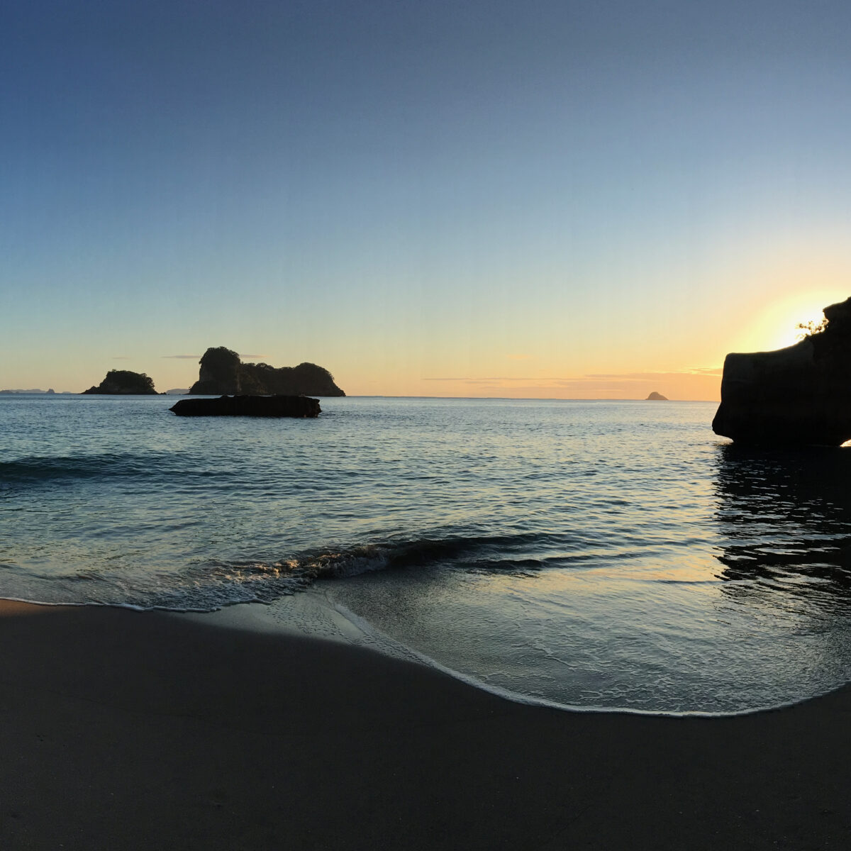 Sunlit crescent beach with calm blue water, towering rock formations, and cliffs, all framed by a sunset glow over the horizon.