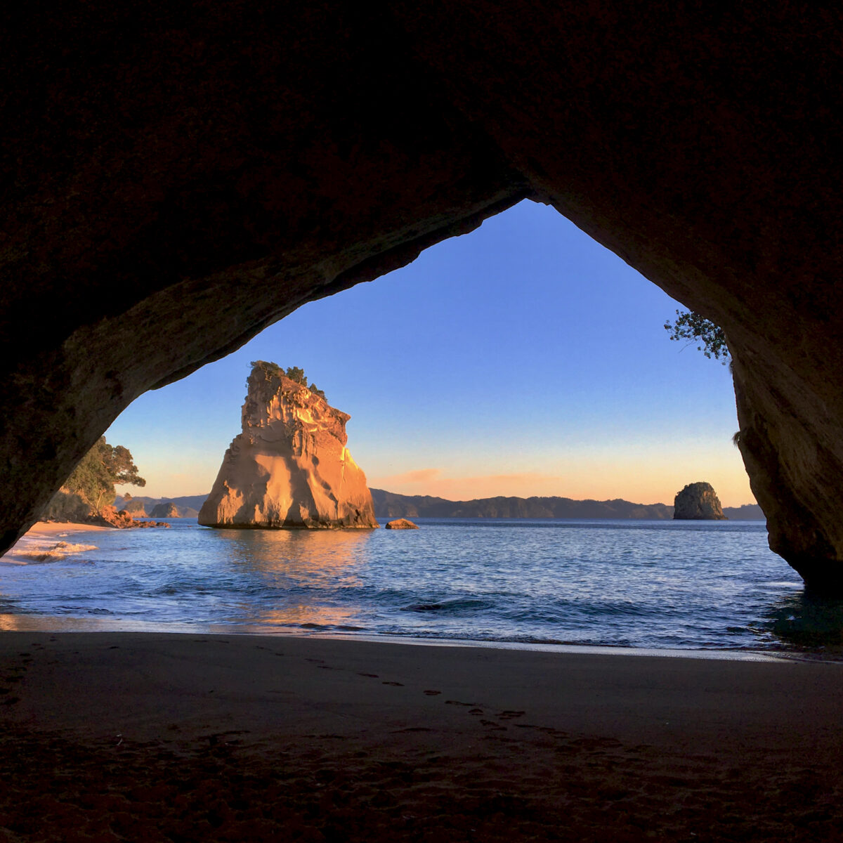 Sunlit sea stacks rise through an opening in a dark rocky cave, with calm water, distant islands, and a clear blue sky.