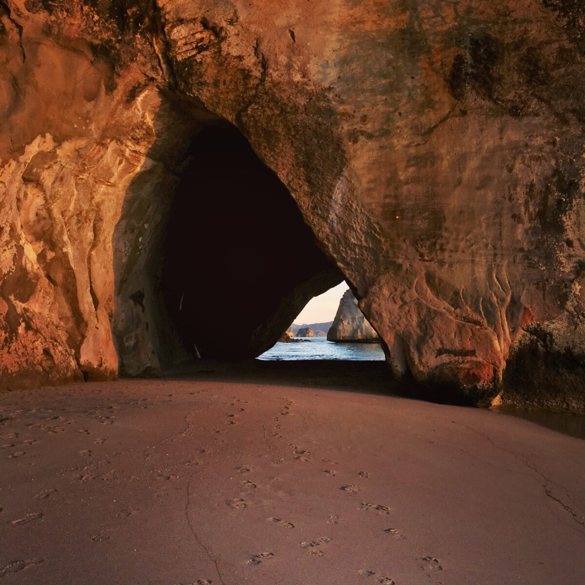 Rocky cave opening in warm sunset light, with sandy beach foreground and distant ocean visible through the arch.