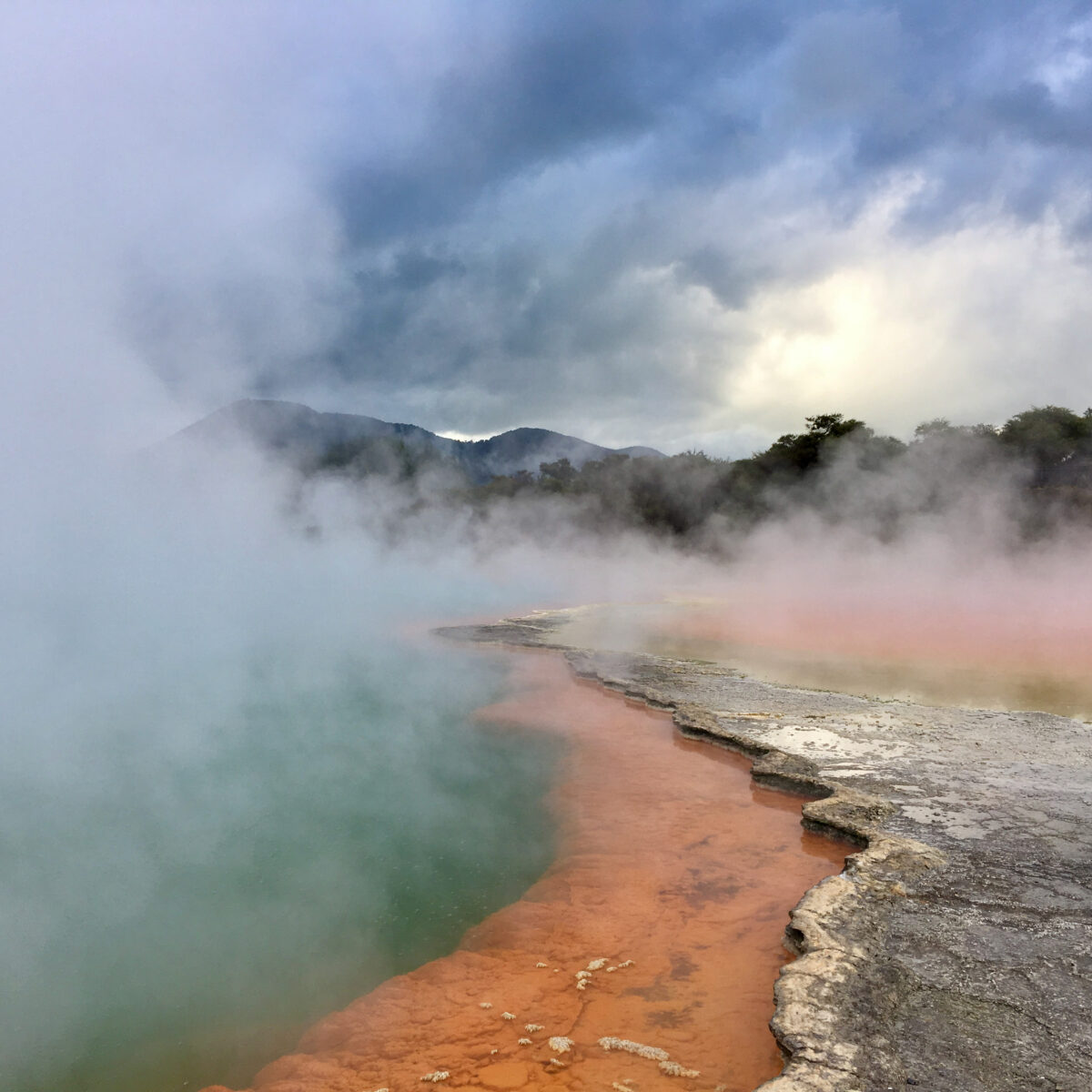 Steam rises from a multicolored hot spring with turquoise water and orange-brown mineral edge, against a gray cloudy sky and distant hills.