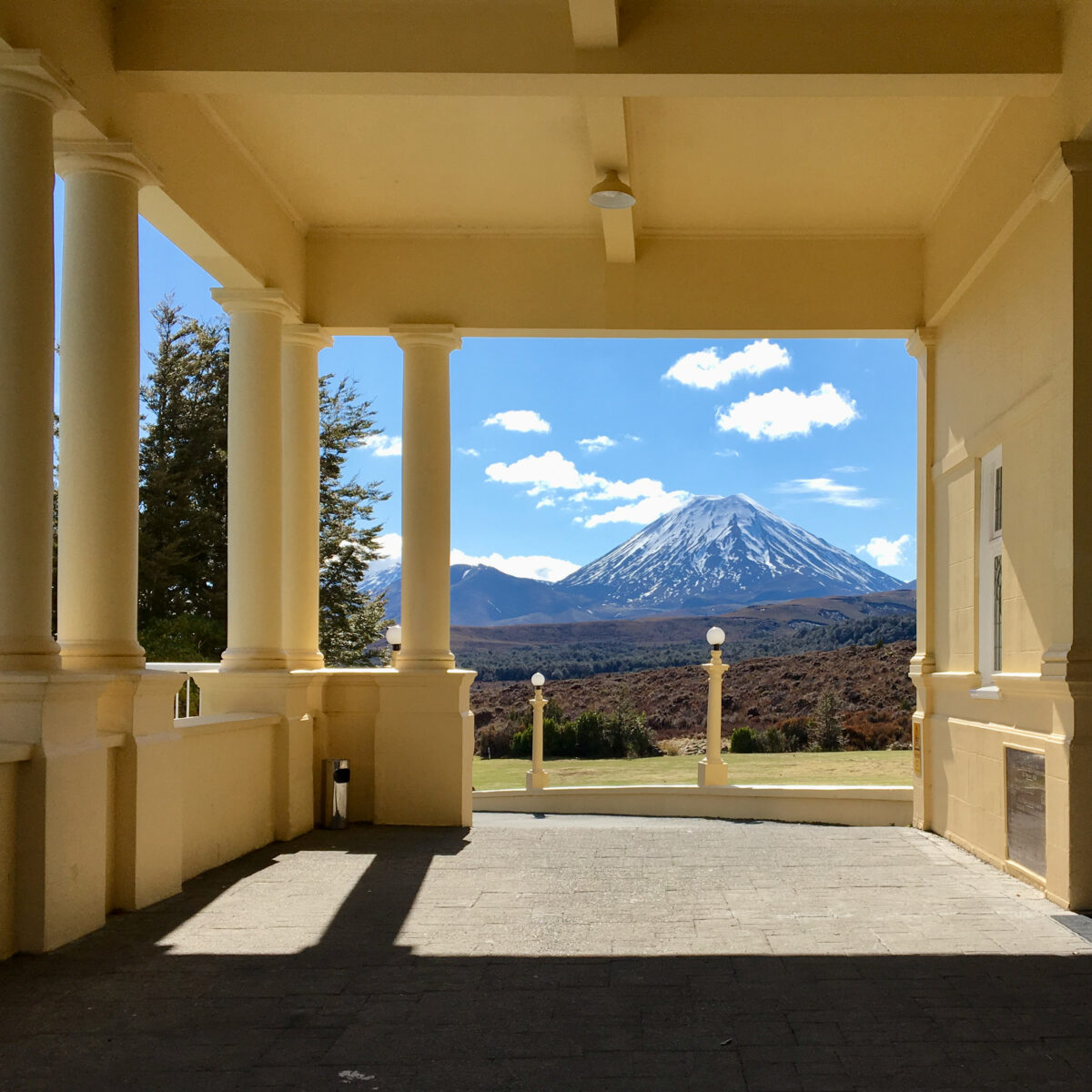 Columns and arches form a beige colonnade framing a view of a snow-capped mountain under a blue sky.