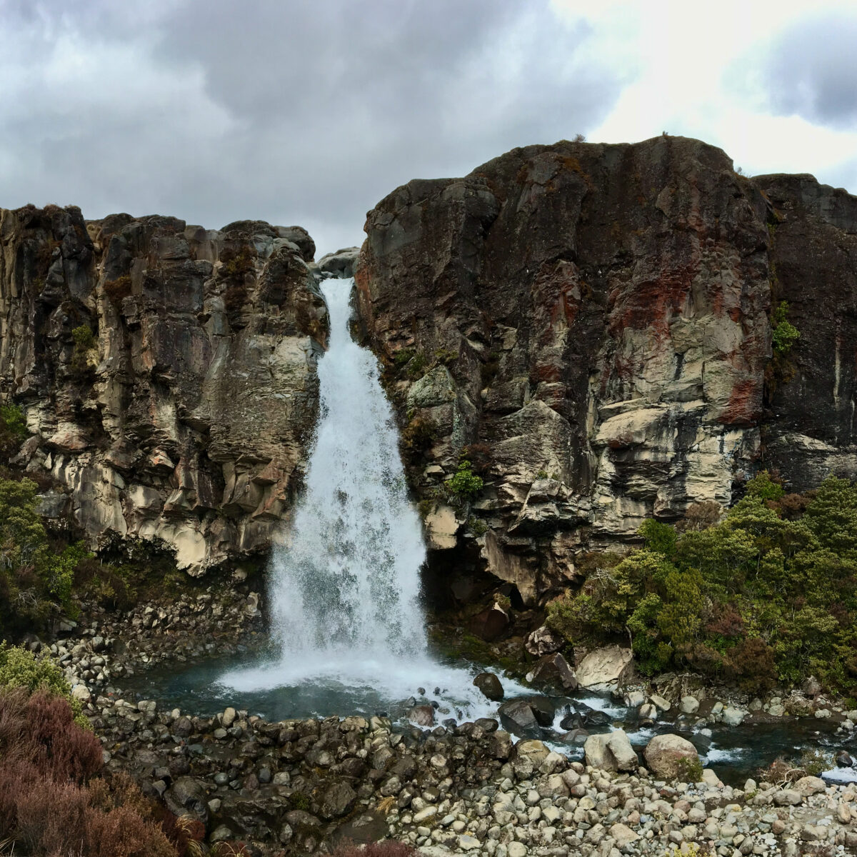A tall waterfall plunges between rugged cliffs into a rocky pool, surrounded by dense green shrubs and brown brush under a cloudy sky.