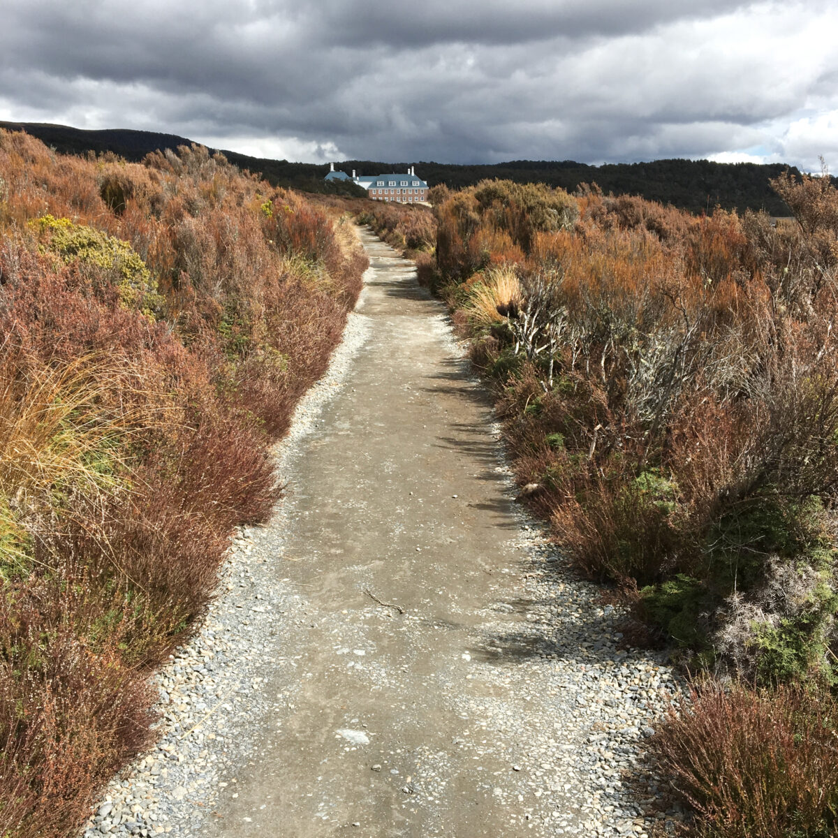 Dirt path through brown shrubs and rocky ground leads toward a distant brick building under dark, cloudy skies.