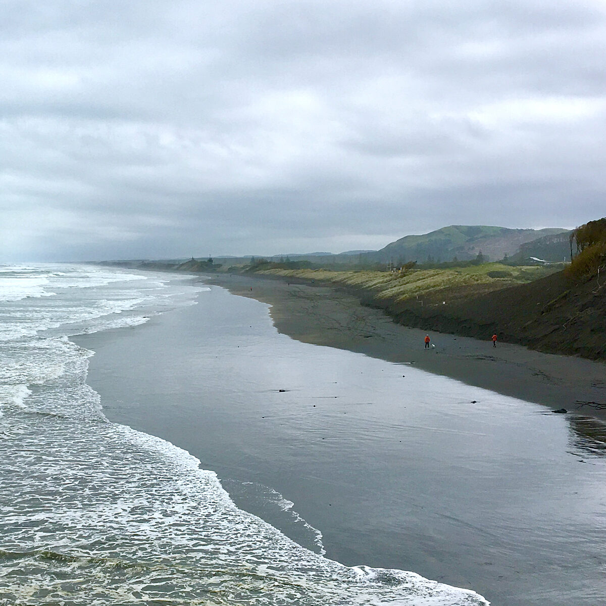 Overcast beach with calm waves, wet sand reflecting gray sky, and distant hills along a rocky coastline.